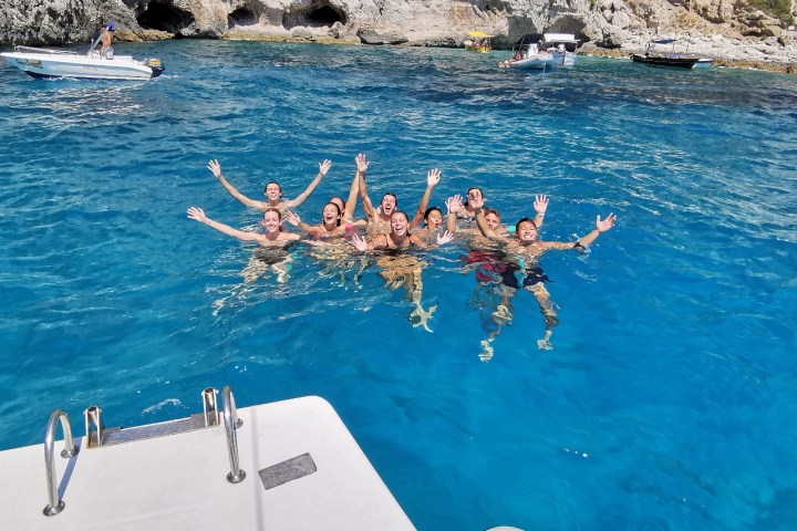 Group of people swimming in clear blue water near rocky cliffs, with a boat platform in the foreground.