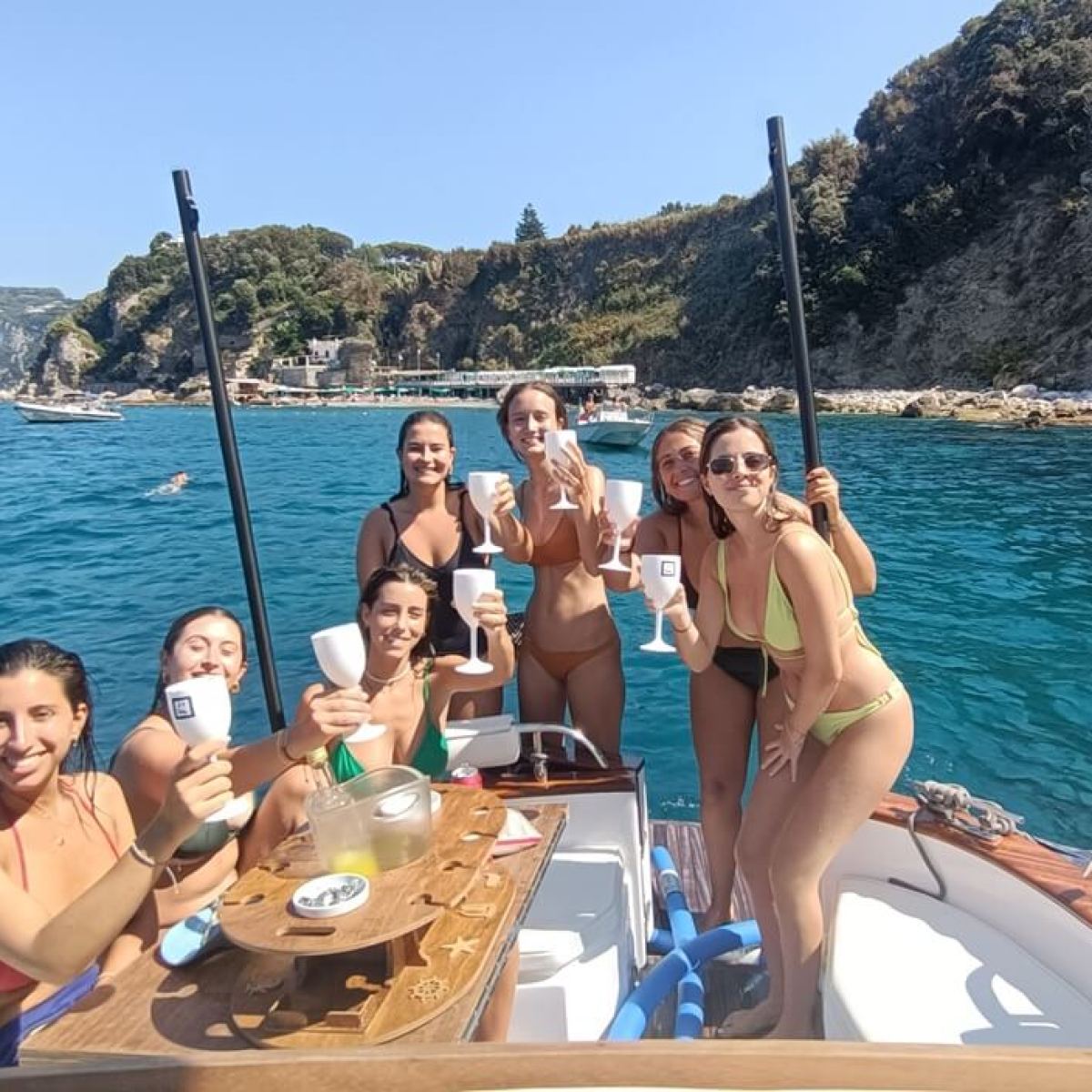 Group of women on a boat, enjoying drinks near a rocky coastline.