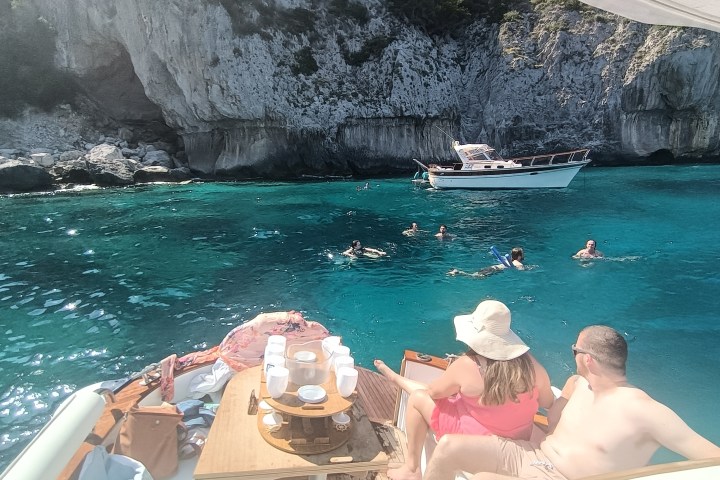 People swimming near cliffs and boats in clear blue water.