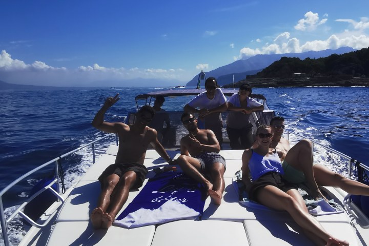 Six people relaxing on a boat in sunny weather with mountains in the background.