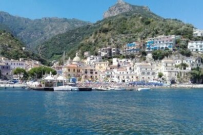Coastal town with houses on hillside, boats docked on calm water, and green mountains in background.