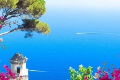 Blue sea view with a small tower, pink flowers, and a tree under a clear sky.