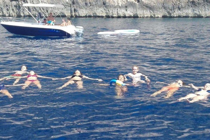 Group of people floating in ocean near boat, rocky coast in background.