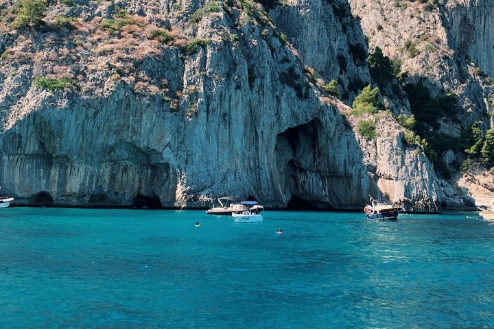 Boats in blue water near rocky cliffs with caves.