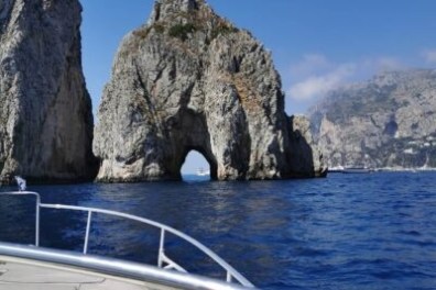 Boat approaching rocky arch formation in blue sea with cliffs in background.