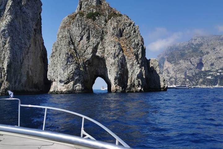 A boat approaching rock arch formations in a blue sea under a clear sky.