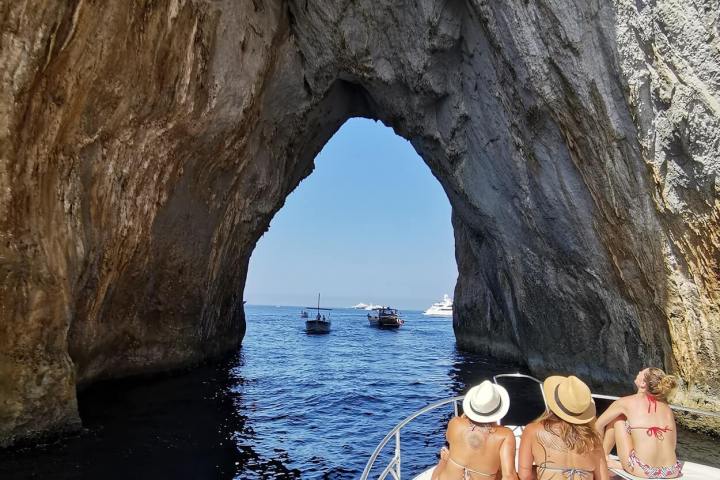 People on a boat approaching a large rock arch over the sea.