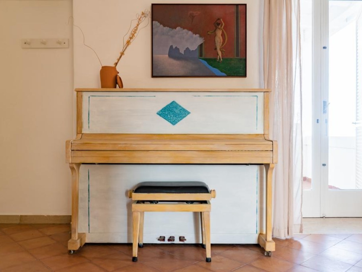 Upright piano with a stool, artwork, and vase on a wall.