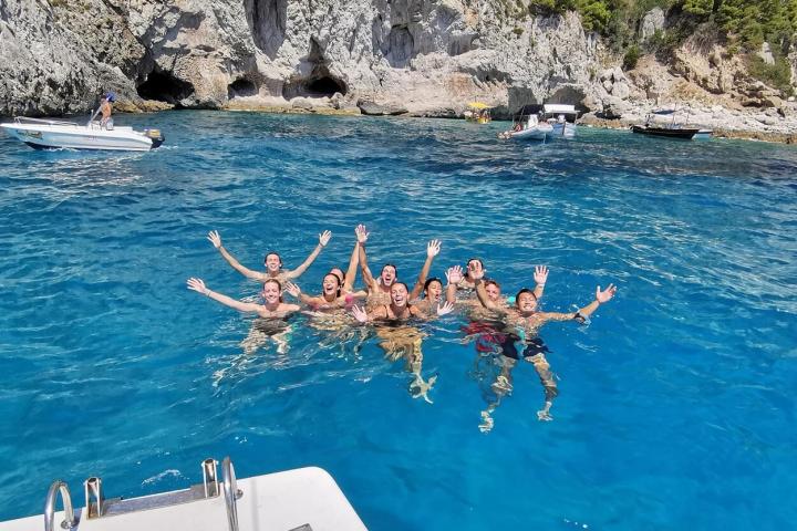 Group of people swimming in clear blue sea, rocky shore in background.